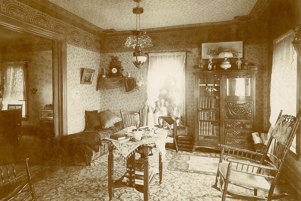 A sepia-toned photograph from the early 1900s shows a Victorian-style room, possibly a living room or parlor. The room features patterned wallpaper, a central table with a lamp, a tall wooden cabinet, and several chairs including rocking chairs.