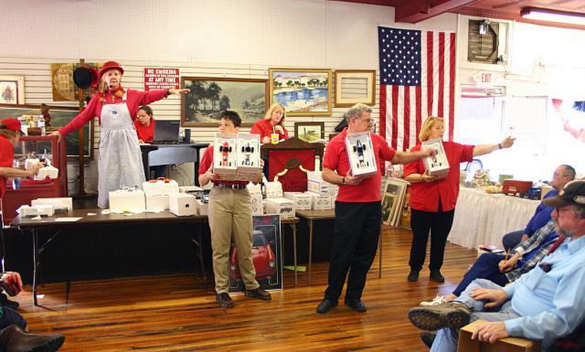 An auction in progress, with an auctioneer and several assistants scanning the crowd for bidders. The auctioneer stands on a stage, while assistants hold up items for sale and point to potential bidders in the audience.