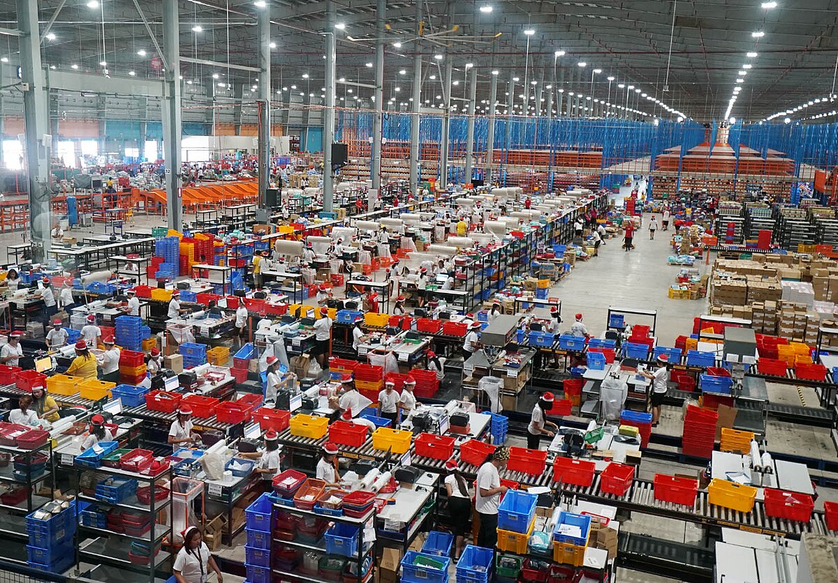 An interior view of a Lazada warehouse in Cabuyao, Laguna, Philippines, in 2018, showing numerous workers sorting and packing items. The warehouse is vast, filled with shelves, conveyor belts, and colorful bins.