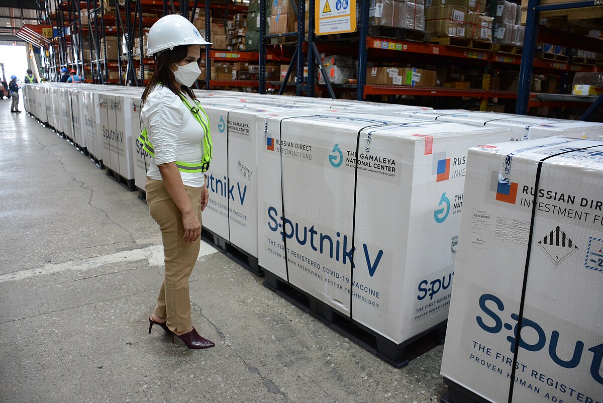 A woman in a hard hat and safety vest stands next to rows of large white boxes labeled "Sputnik V" in a warehouse. These boxes contain shipments of the Russian-made Sputnik V COVID-19 vaccine, an adenoviral vector, in Guatemala in 2021.