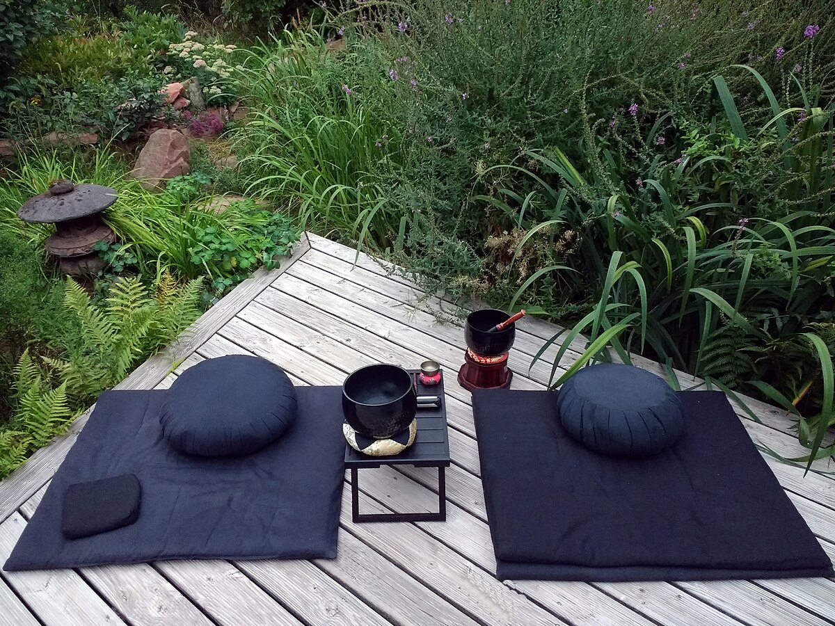 Two zafu meditation cushions and zabuton mats are set up on a wooden deck in a garden for zazen (sitting meditation). Bowls and a small table are placed between the two meditation setups.