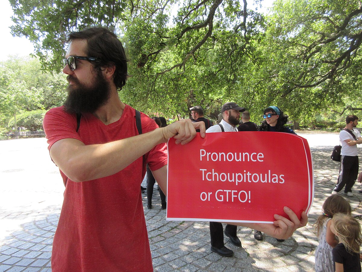 A man with a beard and sunglasses holds a red sign that reads "Pronounce Tchoupitoulas or GTFO!". This sign refers to a shibboleth used by New Orleans residents to identify outsiders during protests against the removal of the Robert E. Lee Monument in 2017.