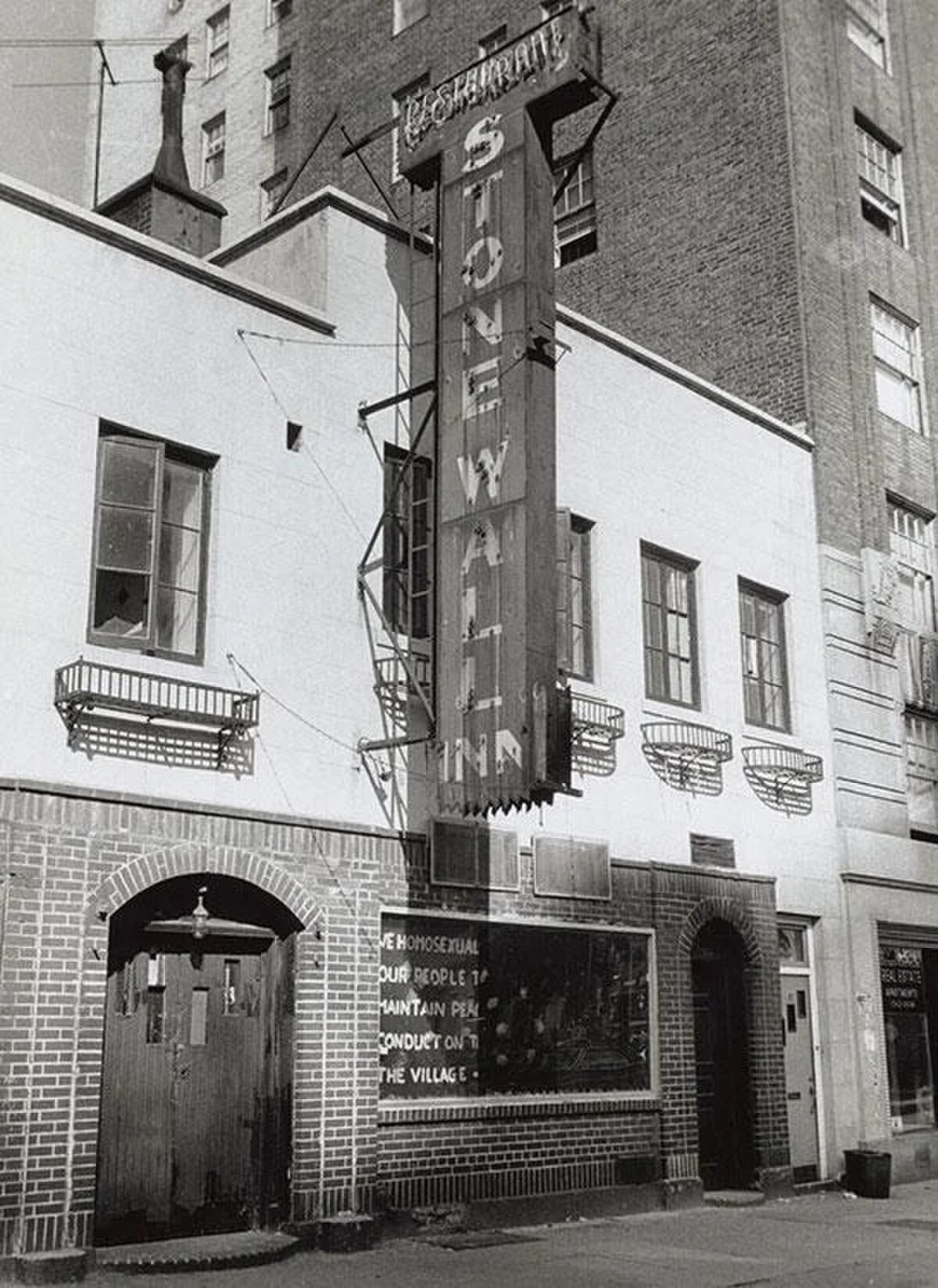 The exterior of the Stonewall Inn, a two-story building with a prominent vertical sign reading "STONEWALL INN RESTAURANT." A banner below the sign displays text, partially visible, that includes "NEW HOMOSEXUAL OUR PEOPLE TO MAINTAIN PEA CONDUCT ON T THE VILLAGE."