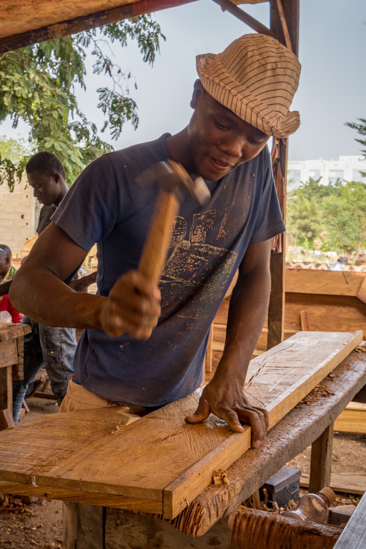 Carpenter in Bamako, Mali, wearing a striped hat and blue t-shirt, hammering a piece of wood. He is working in an open-air workshop with other wooden materials visible in the background.