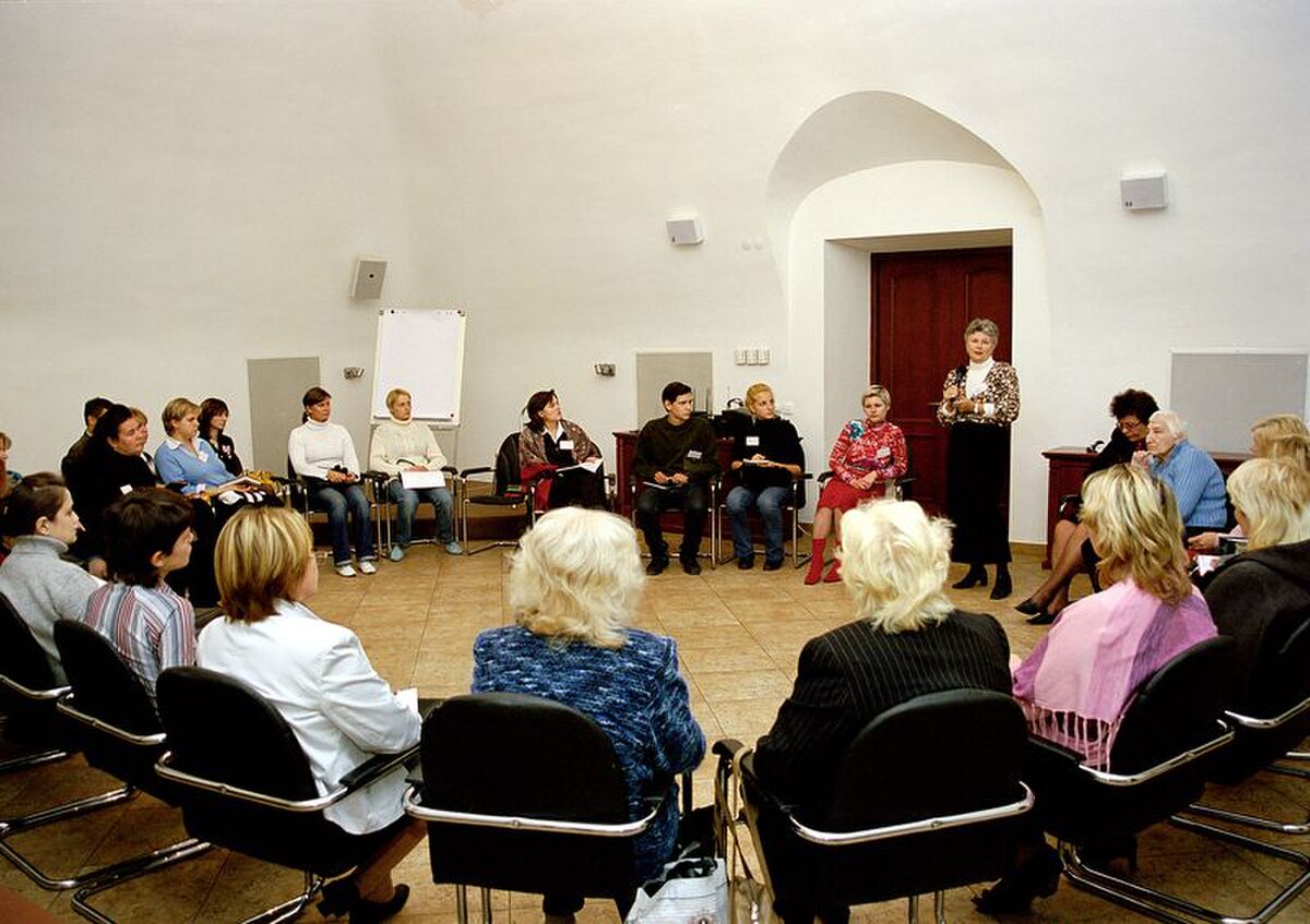 A group therapy session in Ukraine shows a circle of people, mostly women, seated in chairs, with one woman standing and speaking to the group. The room has light-colored walls and a tiled floor.