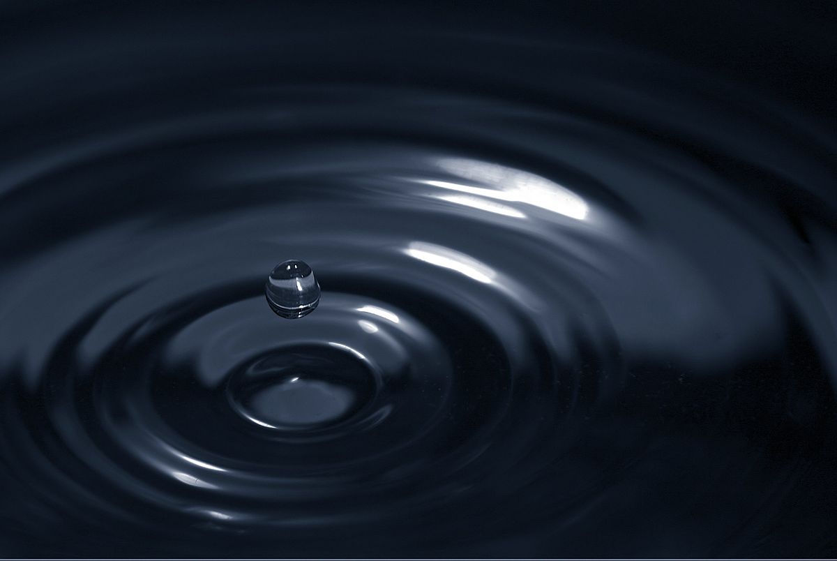 A single water droplet creates ripples on a dark, reflective water surface. The image illustrates the concept of Jivatman (the ripple) being non-different from Brahman (the water).