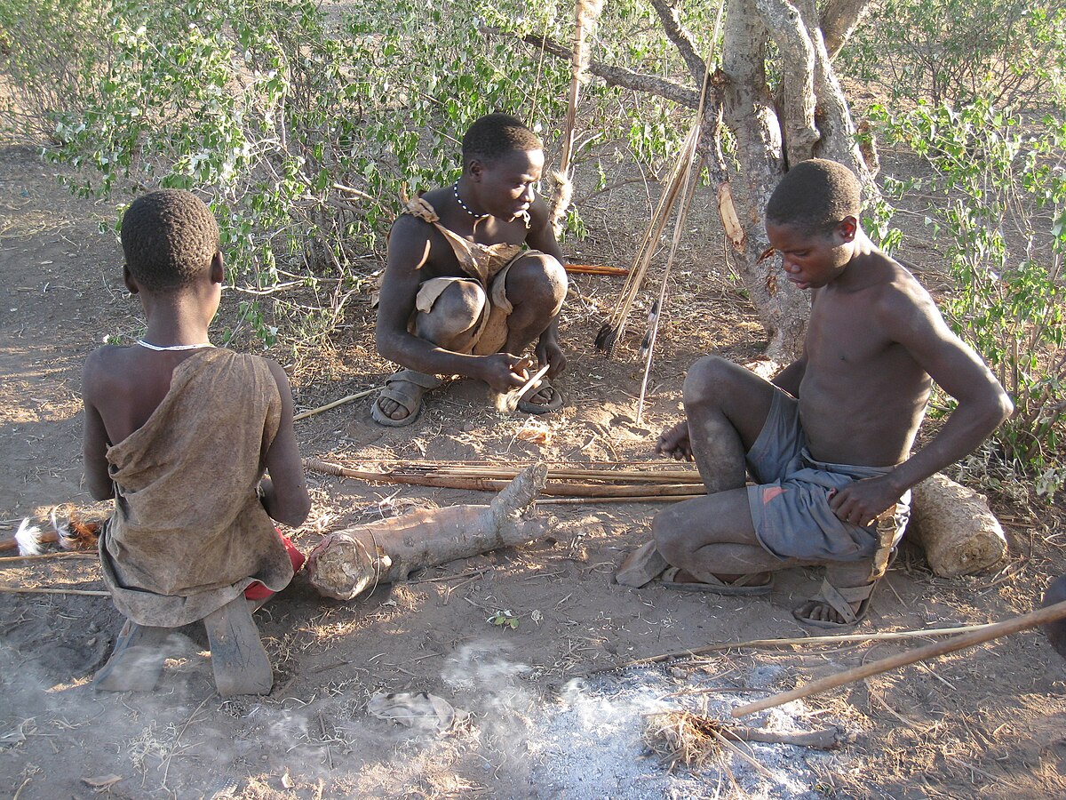 Three Hadza men are gathered around a small fire, engaged in activities such as preparing tools or food. They are dressed in simple attire, and bows and arrows are visible nearby, indicating their hunter-gatherer lifestyle.