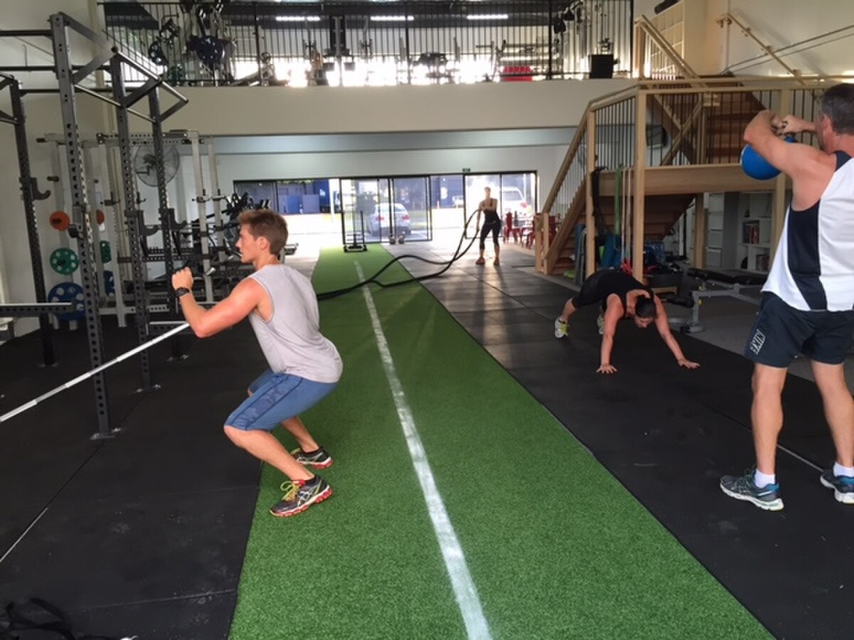 A group of people engaged in a small group fitness session at a gym, performing various exercises. One person is doing squats with a resistance band, another is on the floor in a plank-like position, and a third is swinging a kettlebell, demonstrating strength training activities.