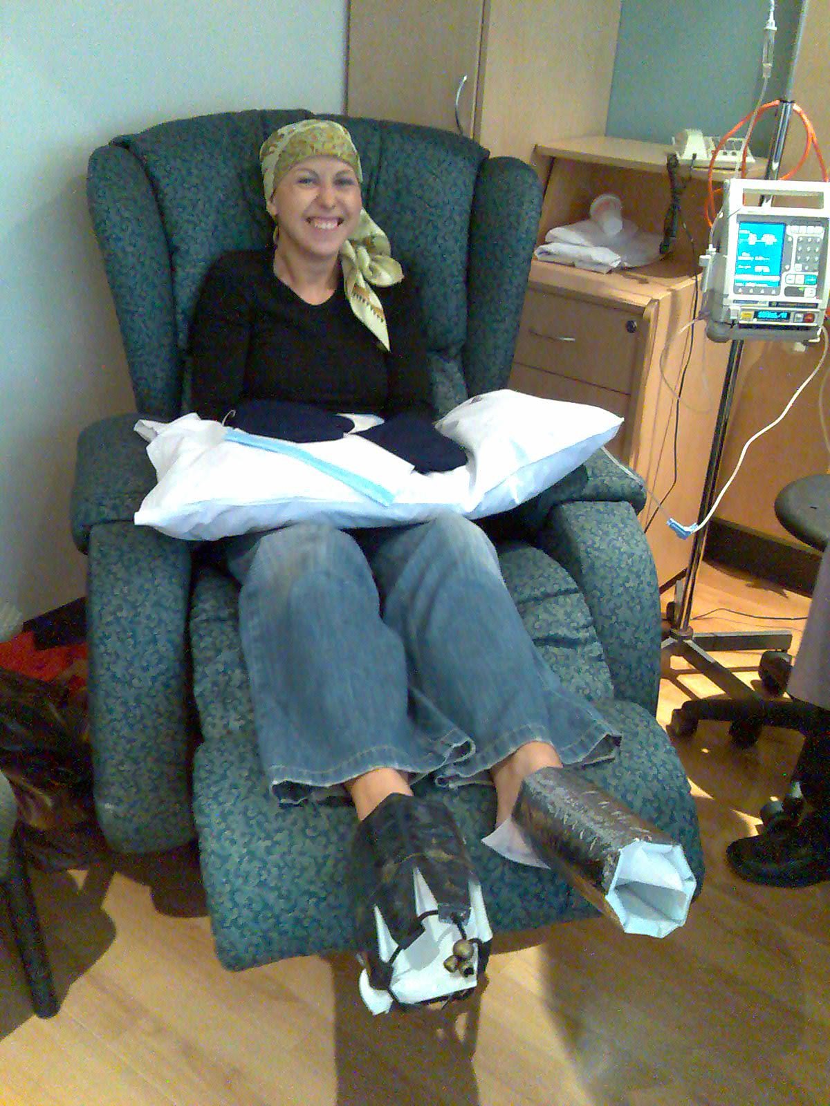 A woman undergoing chemotherapy treatment, seated in a recliner with a headscarf and smiling, while using acral cooling on her hands and feet. Medical equipment, including an IV drip and an infusion pump, is visible next to her.