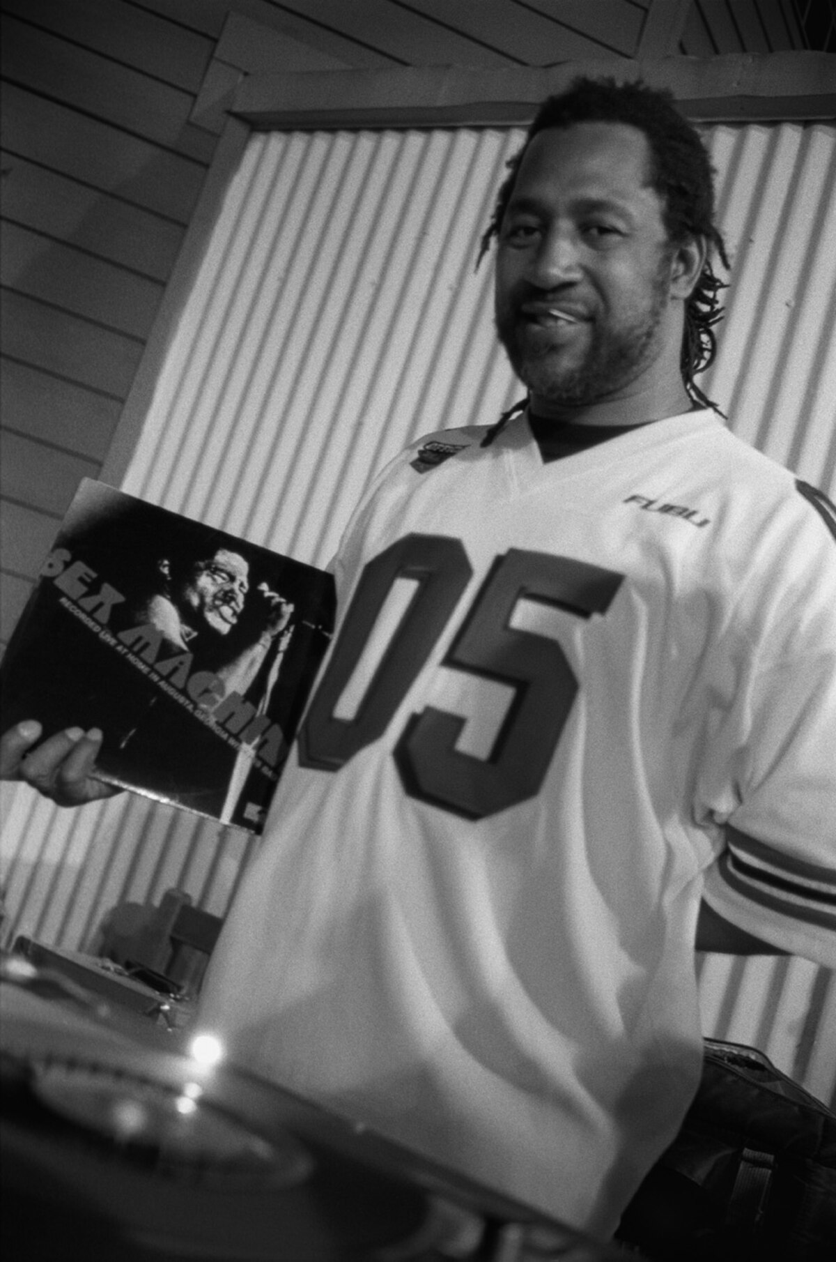 Black and white portrait of DJ Kool Herc in 1999, smiling and holding a vinyl record album. He is wearing a jersey with the number "05" on it.