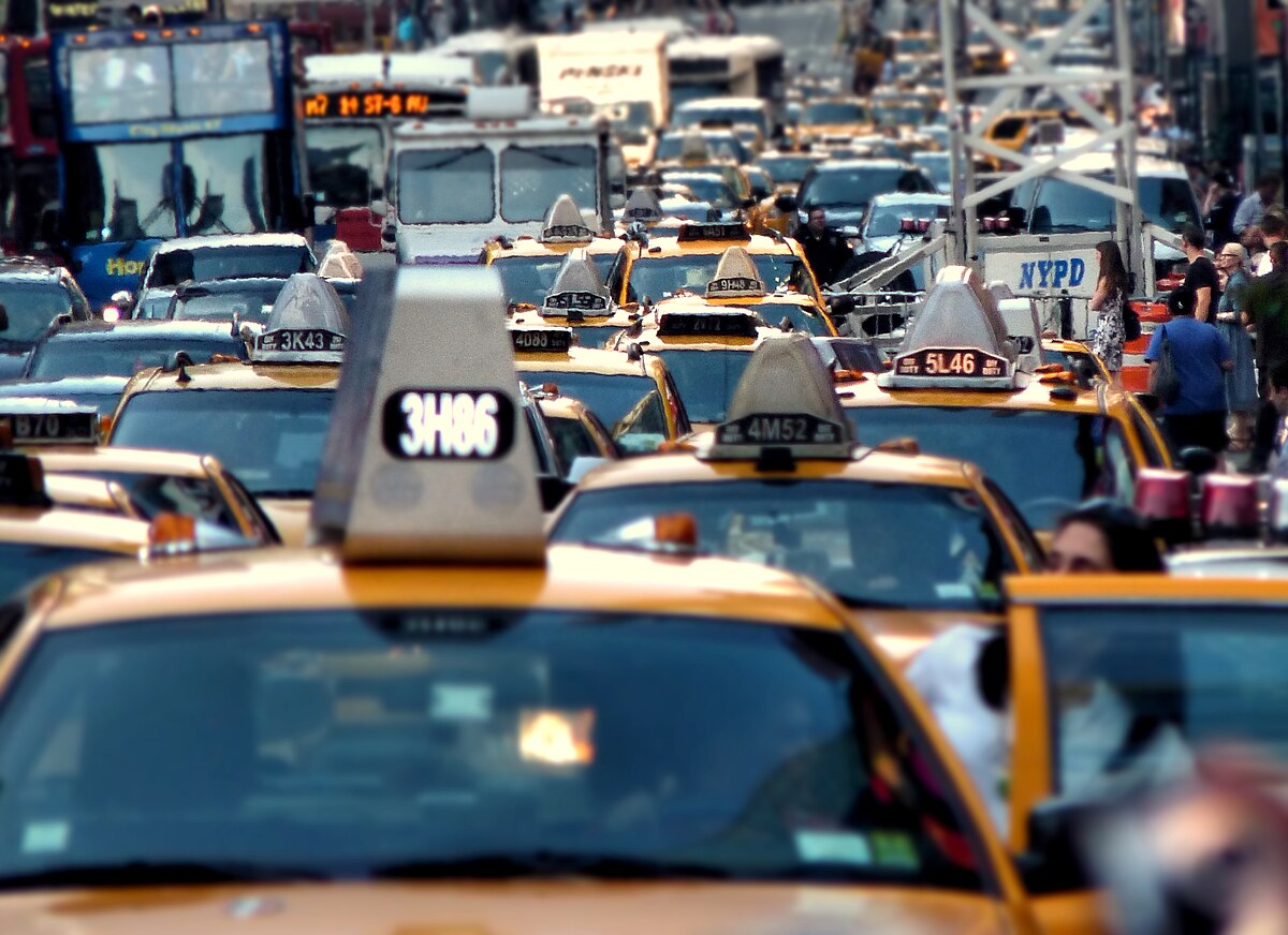 A street-level view of a severe traffic jam in Times Square, Midtown Manhattan, filled with numerous yellow taxi cabs and other vehicles. The image captures the dense congestion with many cars bumper-to-bumper, highlighting the issue of urban traffic.
