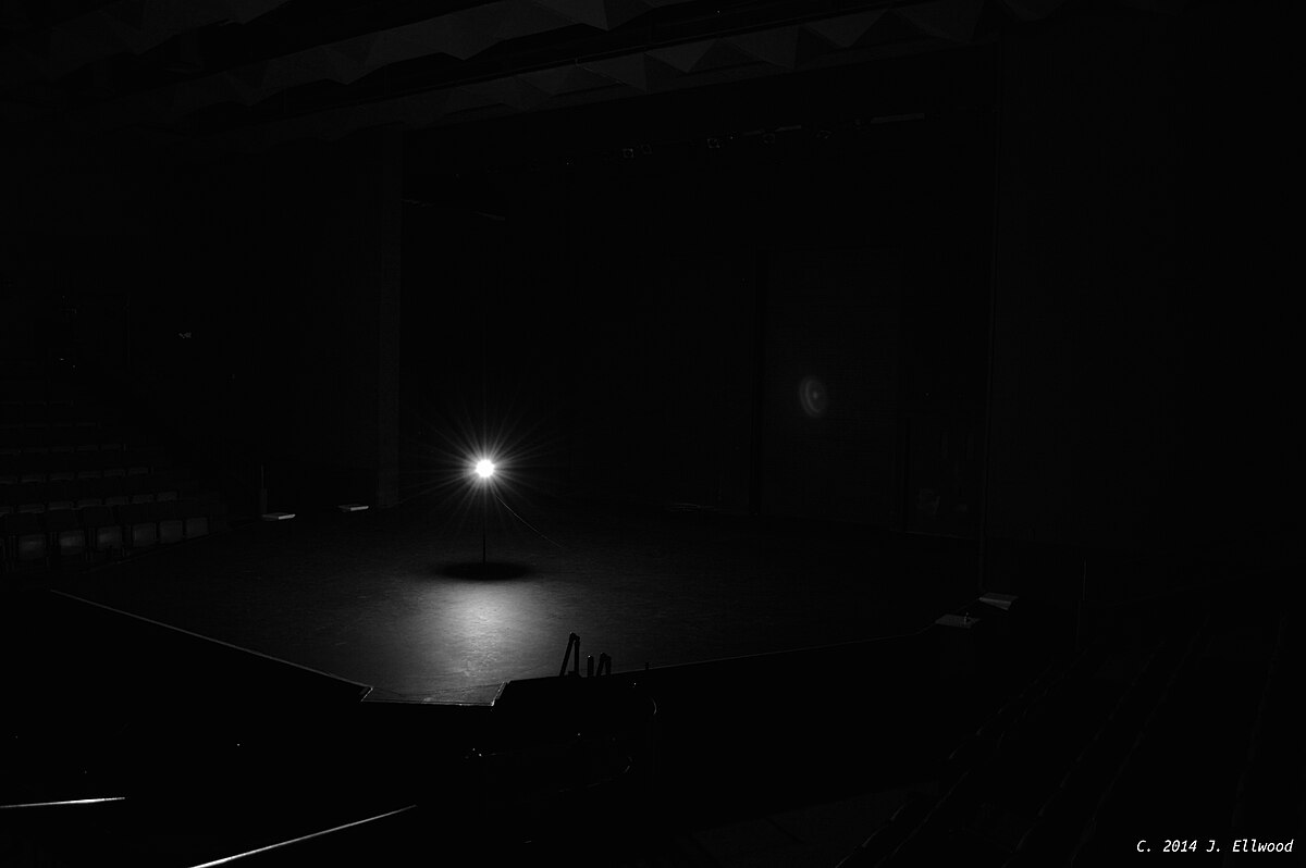A ghost light illuminates an empty stage in a darkened theater, casting a bright circle of light on the center of the stage. The image is in black and white, with a copyright notice "C. 2014 J. Ellwood" in the bottom right corner.