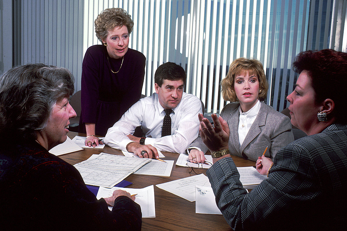 Five individuals are seated around a conference table in an office setting, engaged in a staff meeting. Papers are spread on the table, and participants are actively listening and speaking, demonstrating business communication.