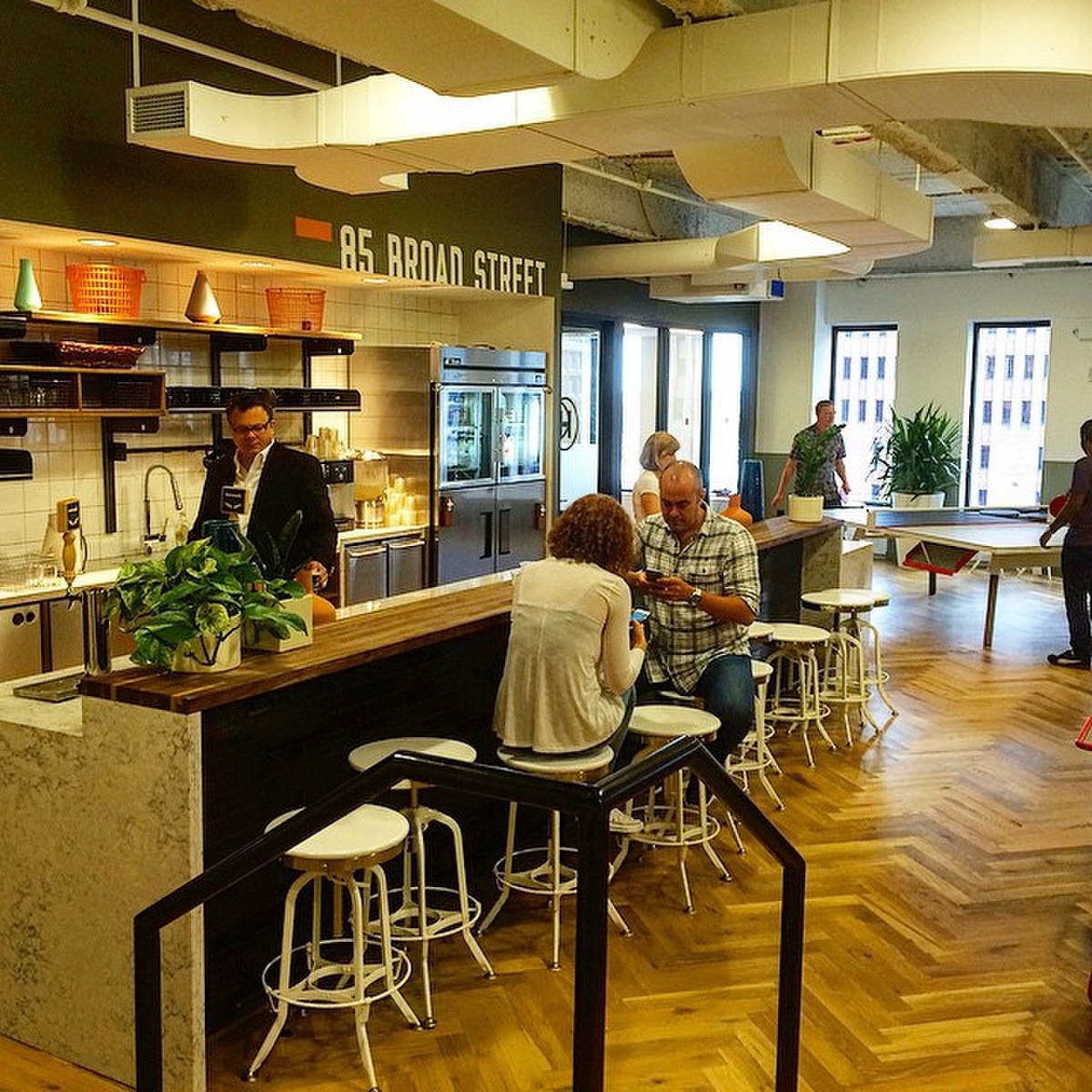 A WeWork kitchen area in New York features a long wooden counter with white bar stools, where several people are seated. The wall above the counter has a sign that reads "85 BROAD STREET", and a ping-pong table is visible in the background.