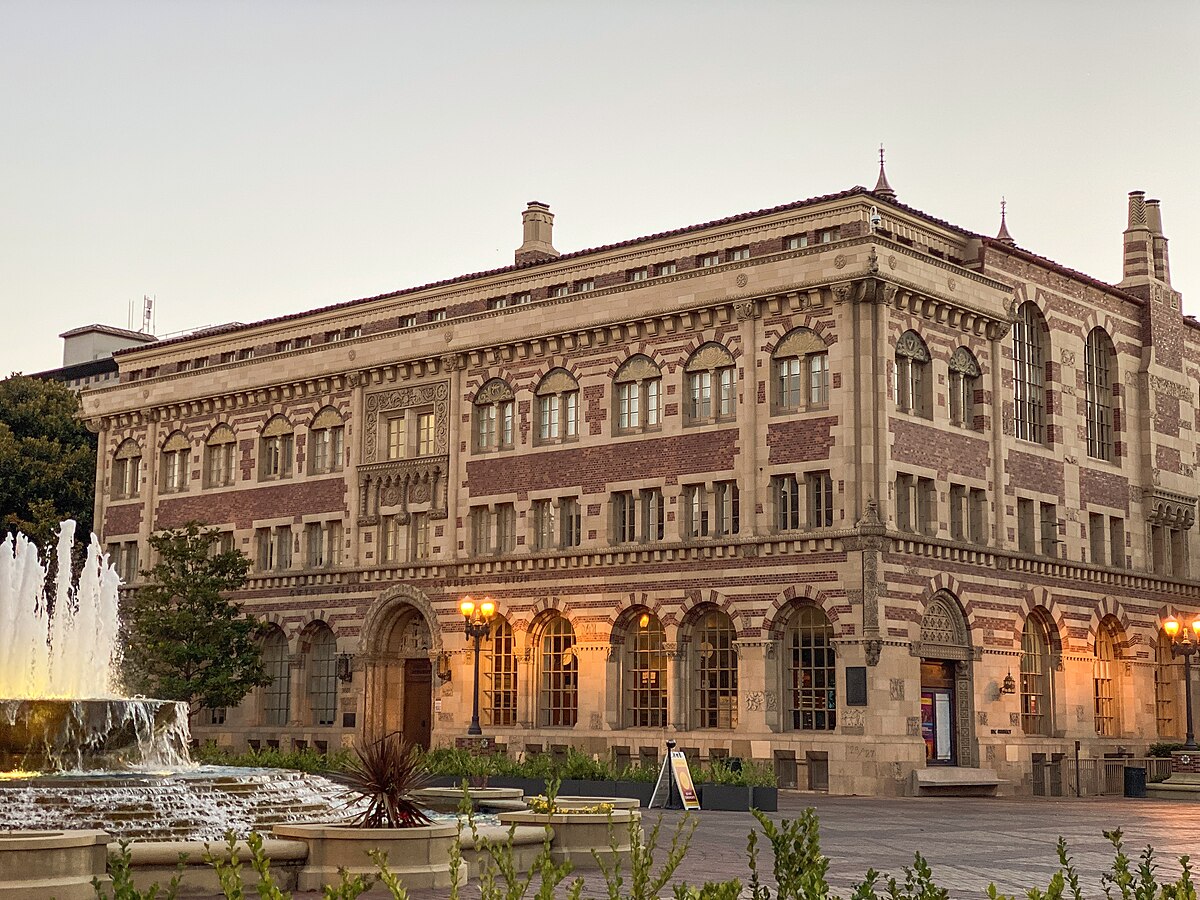 The Gwynn Wilson Student Union on the University Park campus of the University of Southern California is shown with a fountain in the foreground. The building features intricate brickwork and stone detailing, with multiple arched windows and decorative elements.