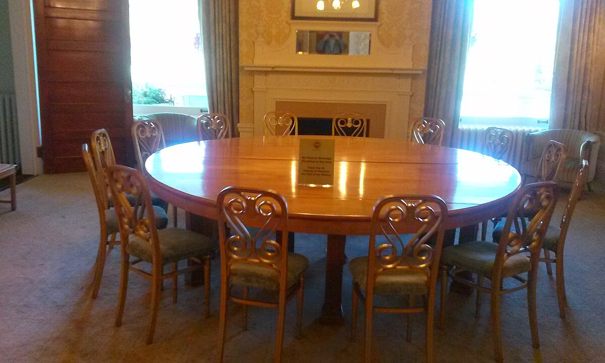 The Gold Room at the Mount Washington Hotel features a large, oval wooden table surrounded by chairs, with a fireplace and mantelpiece in the background. This room is where the International Monetary Fund and World Bank were established.