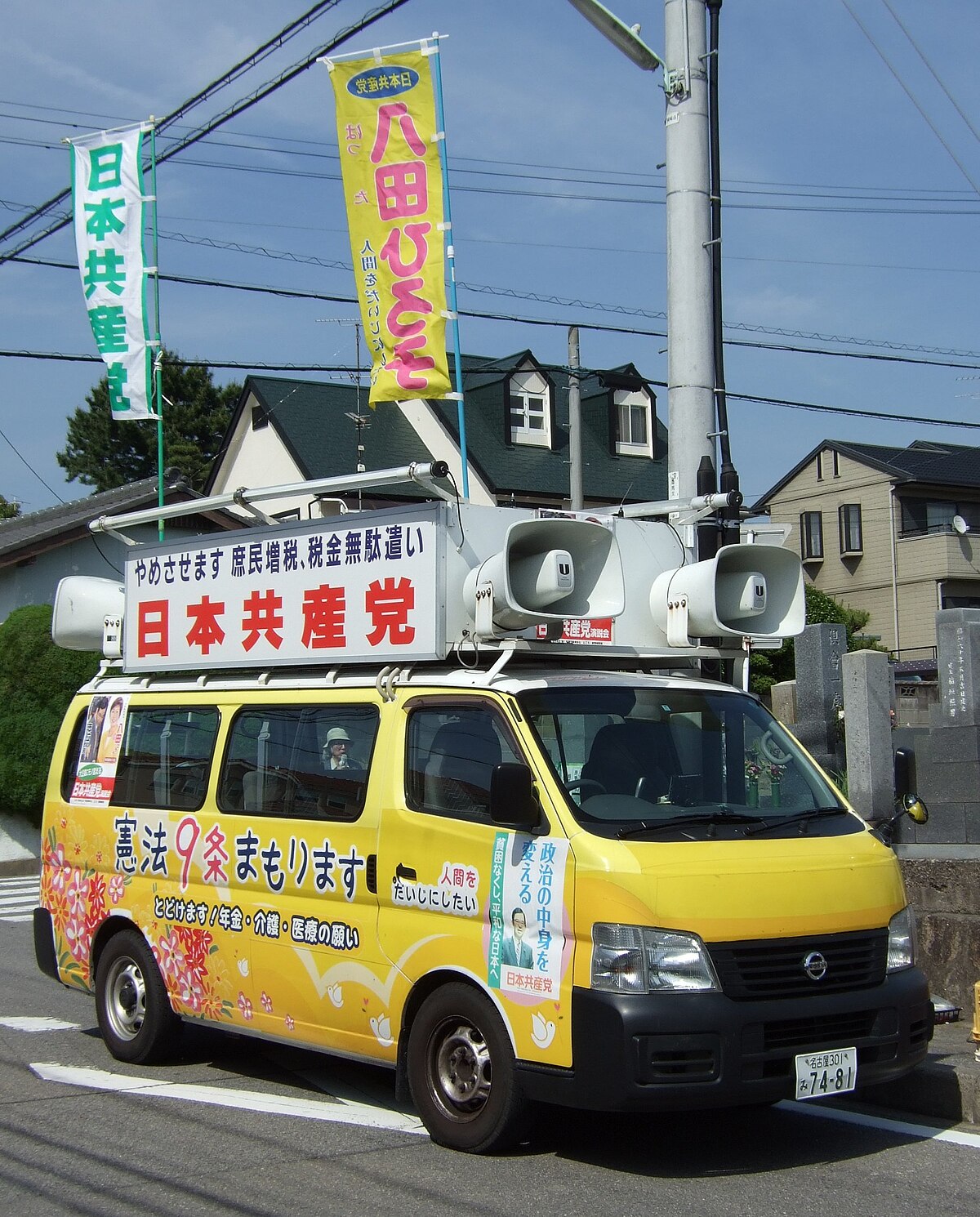 A yellow Nissan Caravan sound truck of the JCP (Japanese Communist Party) parked on a street in Okazaki, Aichi, Japan, on May 20, 2009. The truck is adorned with banners and signs in Japanese, featuring large loudspeakers on its roof and political messages on its sides.