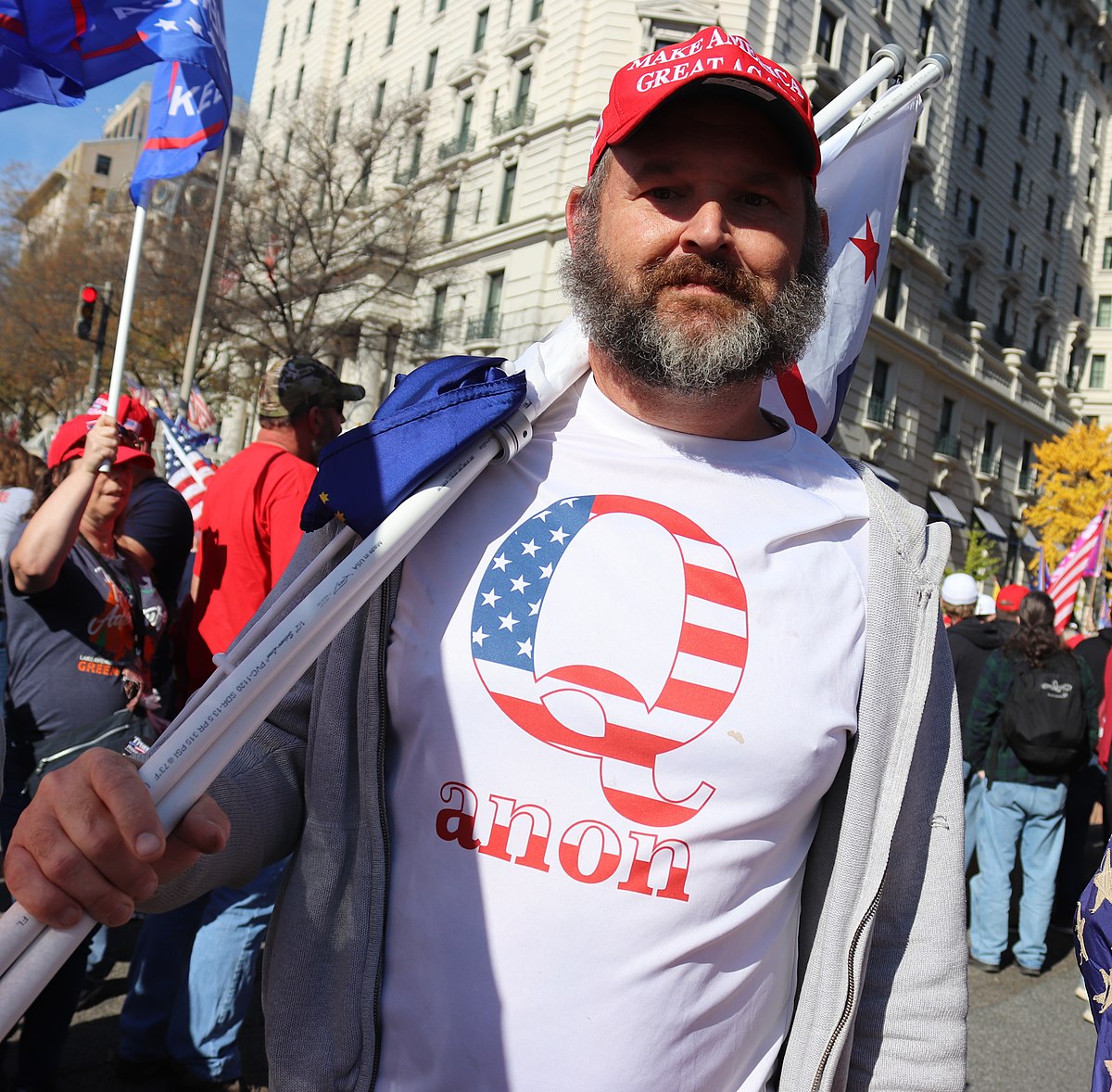 A pro-Trump protester wears a white t-shirt with a QAnon logo featuring a 'Q' in red and white stripes and blue stars, with the word "anon" below it. He is also wearing a red "MAKE AMERICA GREAT AGAIN" hat.