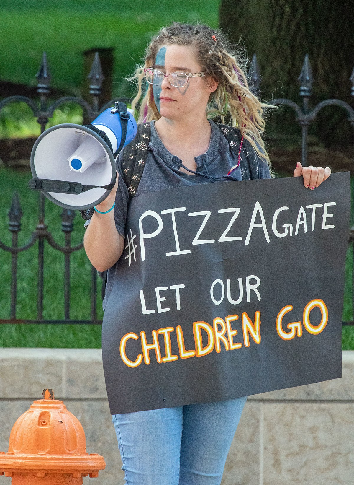 A protester holds a sign that reads "#PIZZAGATE LET OUR CHILDREN GO" in black, white, and orange lettering. The protester has blue and white face paint and is holding a megaphone.