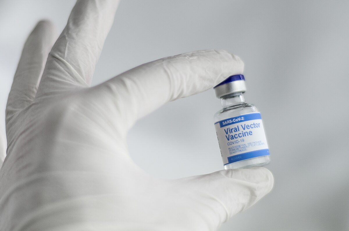 A gloved hand holds a small glass vial labeled "Viral Vector Vaccine" and "COVID-19." The vial has a blue cap and contains a clear liquid.