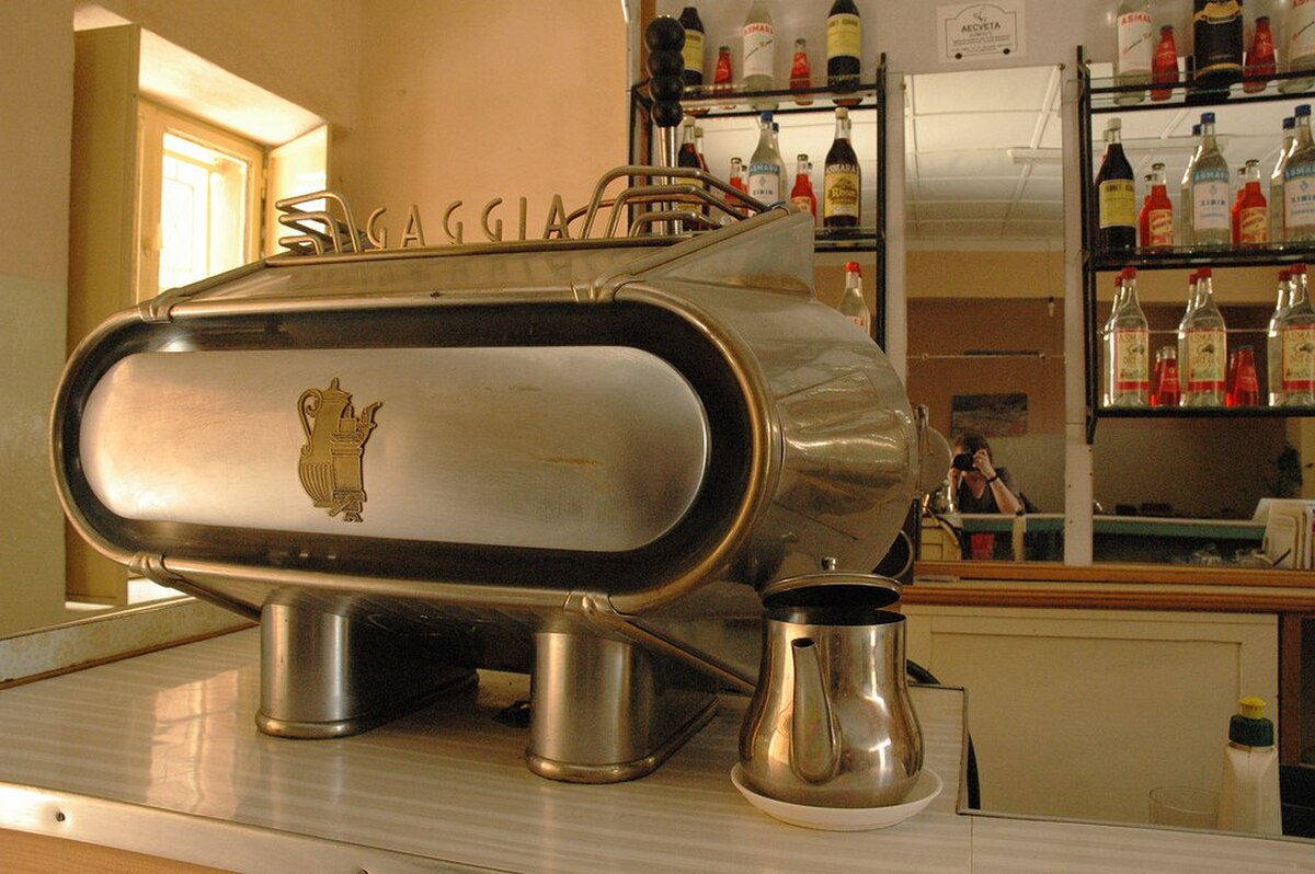A vintage Gaggia espresso machine is displayed on a counter, featuring a polished metal exterior with the "GAGGIA" logo and an embossed coffee pot design. Behind the machine, shelves are visible with various bottles, suggesting a bar or cafe setting. A small metal pitcher sits on the counter next to the machine.
