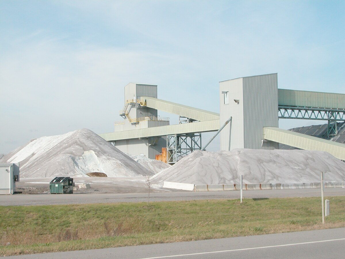An industrial salt mine facility is shown with large piles of extracted salt next to processing buildings and conveyor systems. The scene is set under a clear sky, with a road in the foreground.