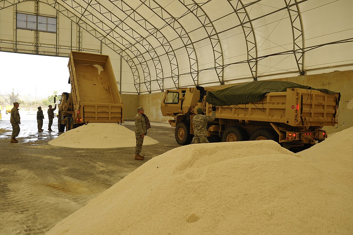 Large mounds of road salt are stored inside a large arched structure, with two dump trucks actively unloading more salt. Several individuals in military-style uniforms are present, overseeing the operation.
