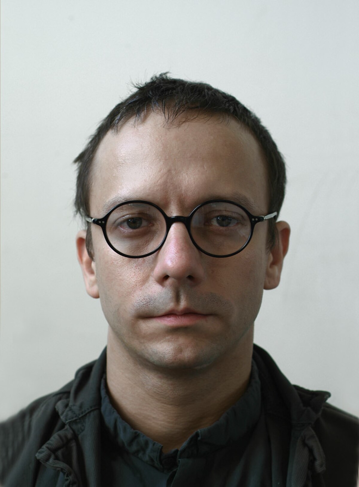 Portrait of Alex Dodge, featuring his natural, unedited face. He is wearing round glasses and a dark jacket, with a neutral expression.