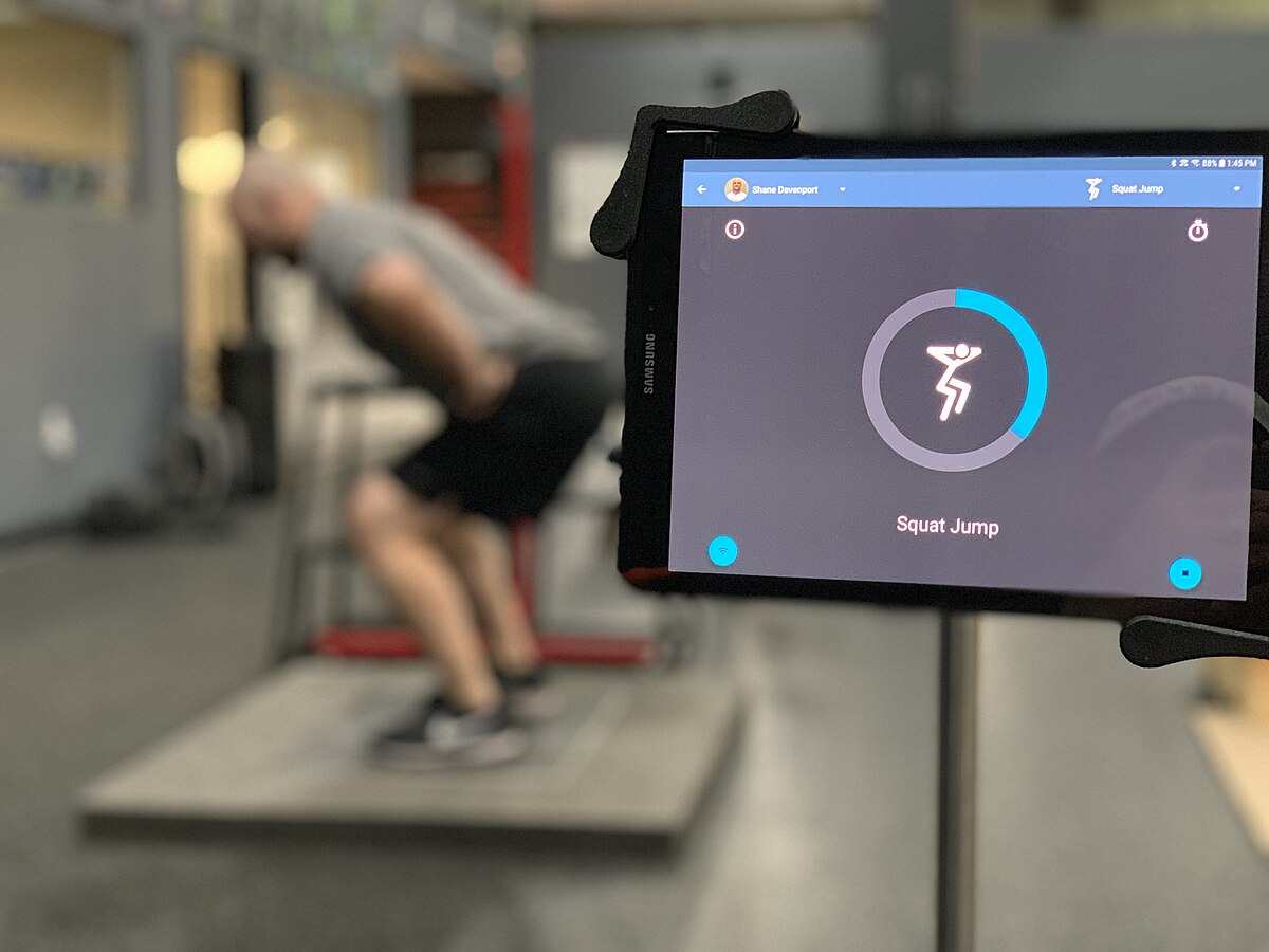 A person performs a squat jump on Hawkin Dynamics force plates, with a tablet in the foreground displaying a "Squat Jump" application interface. The tablet shows a circular progress indicator and a stylized icon of a person squatting.