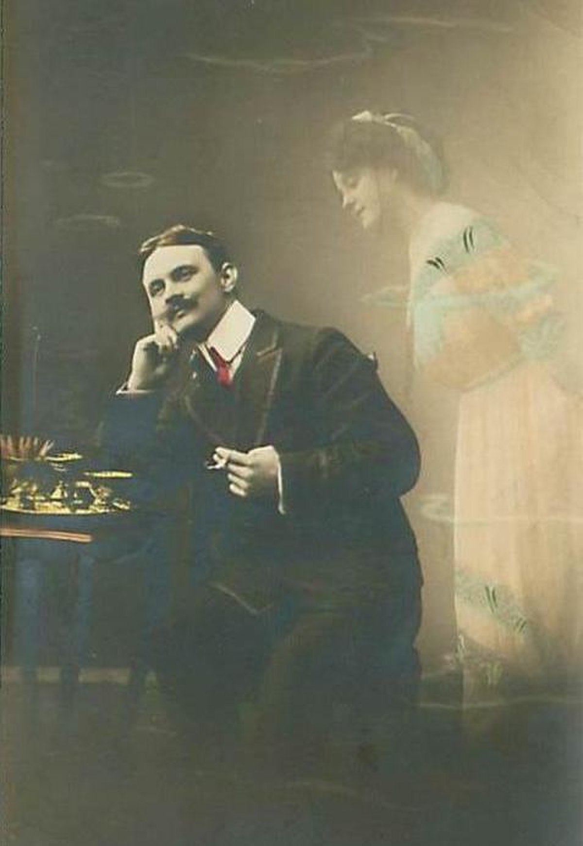 A 1912 postcard illustrates the concept of a man daydreaming about a woman who is not present, showing a seated man with a mustache, looking thoughtfully into the distance, while a translucent, ethereal image of a woman appears to his right. The man is dressed in a suit with a red tie, and the woman is in a light-colored dress.
