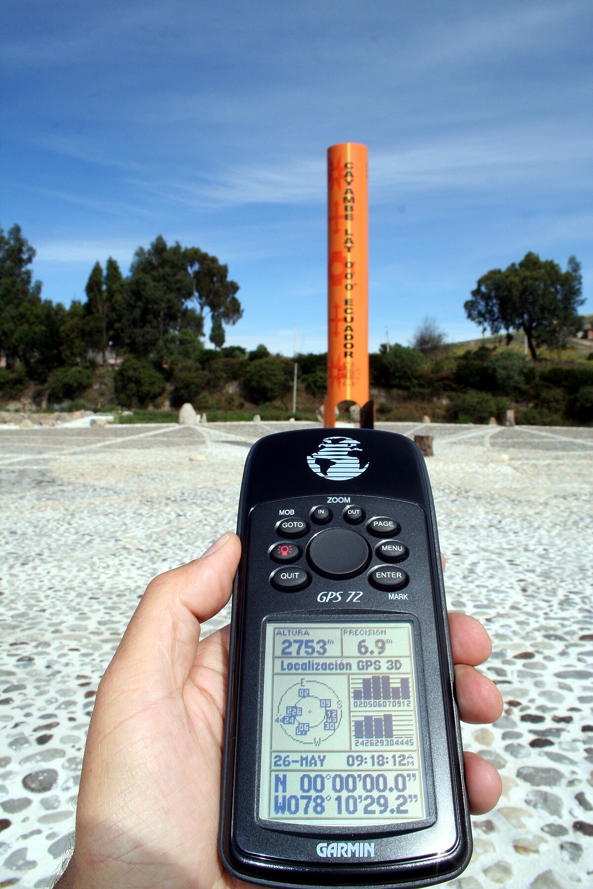 A hand holds a Garmin GPS 72 device displaying coordinates N 00°00'00.0" W 078°10'29.2", indicating the equator, with an orange monument in the background. The GPS screen also shows Altitude 2753 m, Speed 6.9 km/h, and "Localizacion GPS 3D".