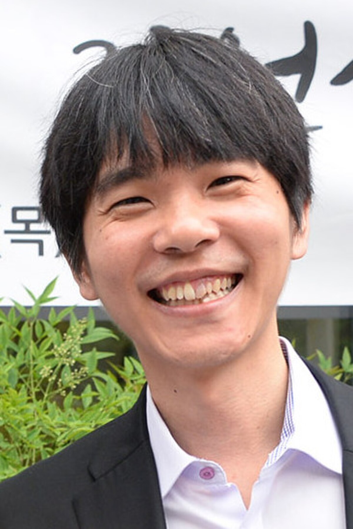 Portrait of Lee Sedol, a South Korean professional Go player, smiling at the camera. He has dark hair and is wearing a dark suit jacket over a light-colored shirt.