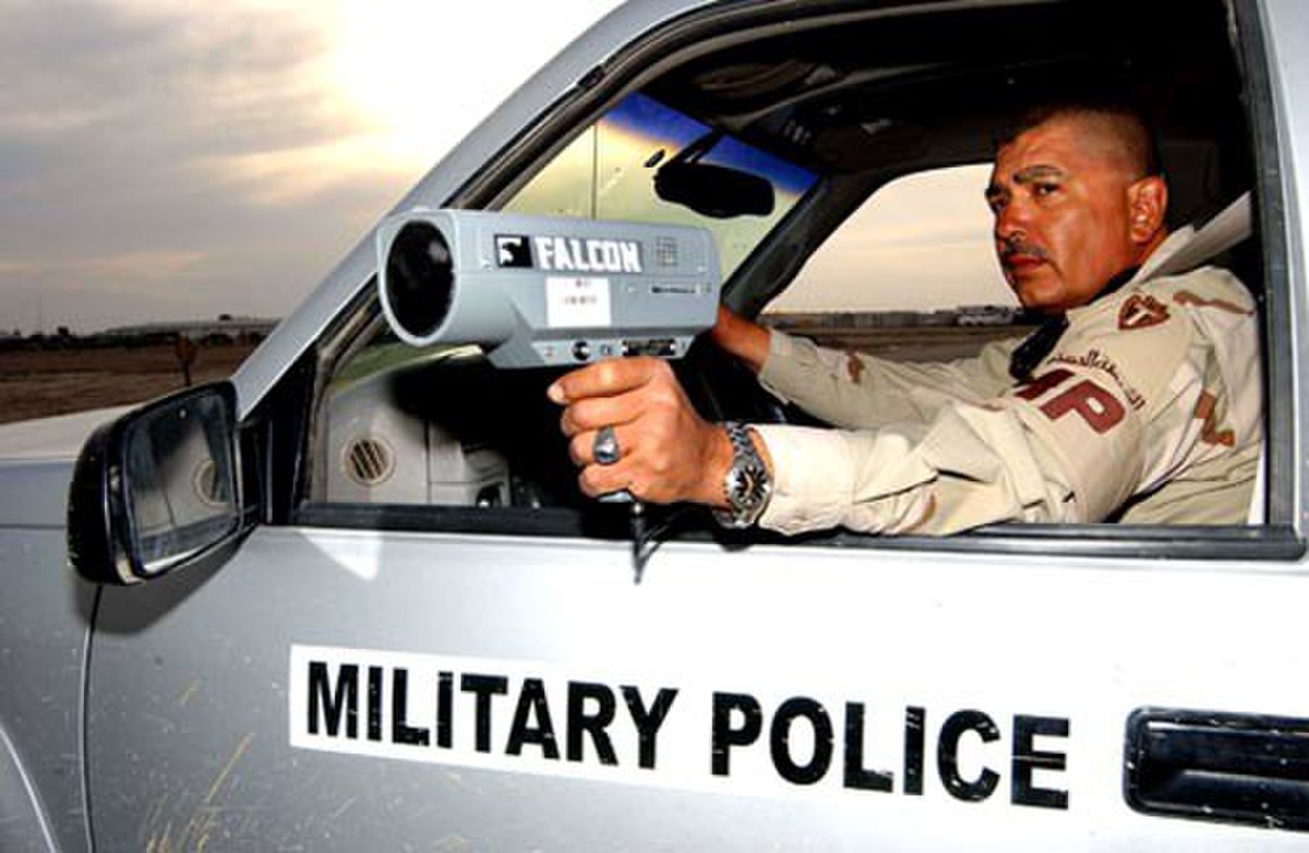 A U.S. Military Police officer uses a Falcon radar gun from inside a vehicle. The radar gun, labeled "FALCON", is pointed out the window, demonstrating an application of the Doppler effect for speed detection.