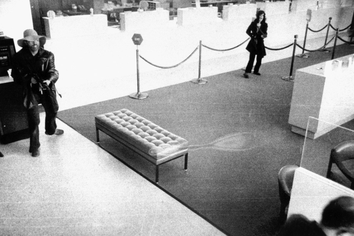 A black and white image shows a bank robbery in progress, with two individuals, one holding a weapon, moving through a bank lobby. The scene depicts the bank robbery by Patty Hearst and Symbionese Liberation Army members.
