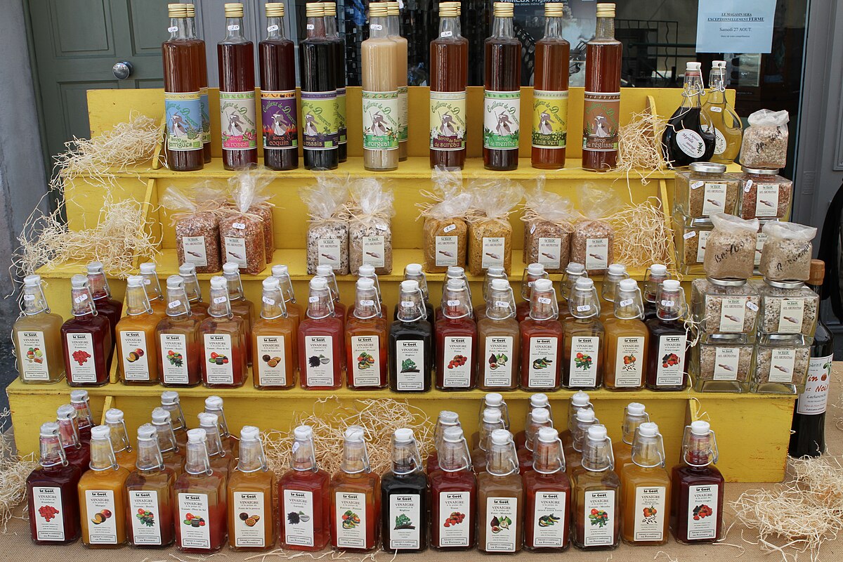 A display of various flavored vinegars for culinary use, on sale in France. The bottles are arranged on yellow tiered shelves, with some small jars of other products also visible.