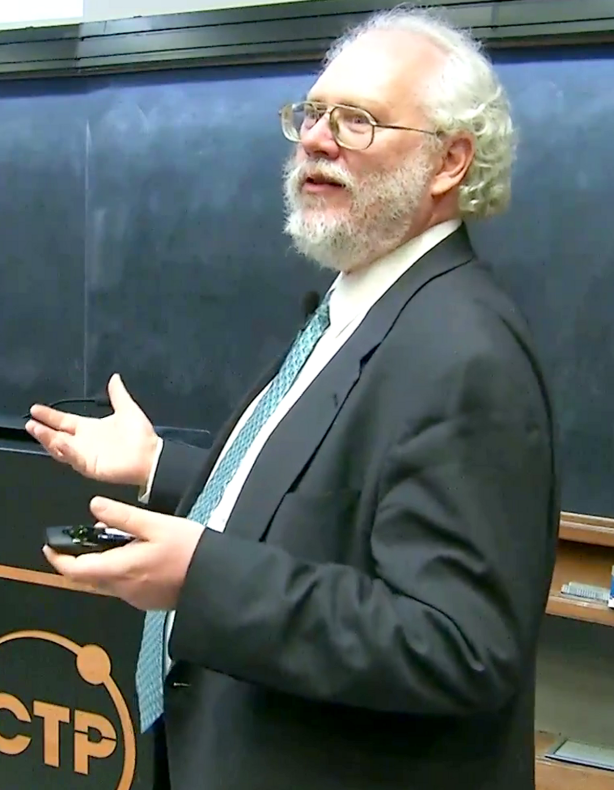 Peter Shor is shown speaking at the 2017 Dirac Medal Award Ceremony, gesturing with his hands. He is a man with a white beard and glasses, wearing a suit and tie, standing in front of a dark chalkboard.