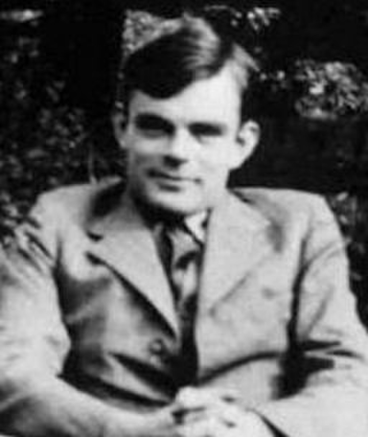 Portrait of Alan Turing, a British mathematician and computer scientist, shown seated outdoors. He is wearing a suit jacket and tie, looking directly at the camera.