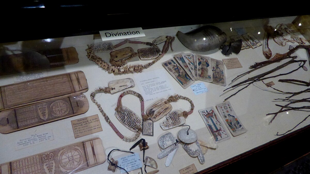 A display on divination at the Pitt Rivers Museum in Oxford, England, showcasing a variety of cross-cultural items used for fortune-telling. Objects include carved wooden tablets, bone or shell pieces, playing cards, and natural materials like branches.