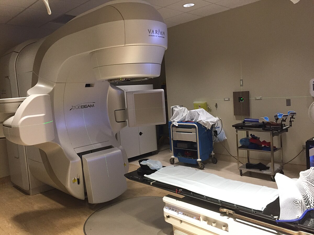 A Varian TrueBeam Linear Accelerator, a large medical device used for delivering Intensity-Modulated Radiation Therapy (IMRT), is shown in a treatment room with a patient bed.