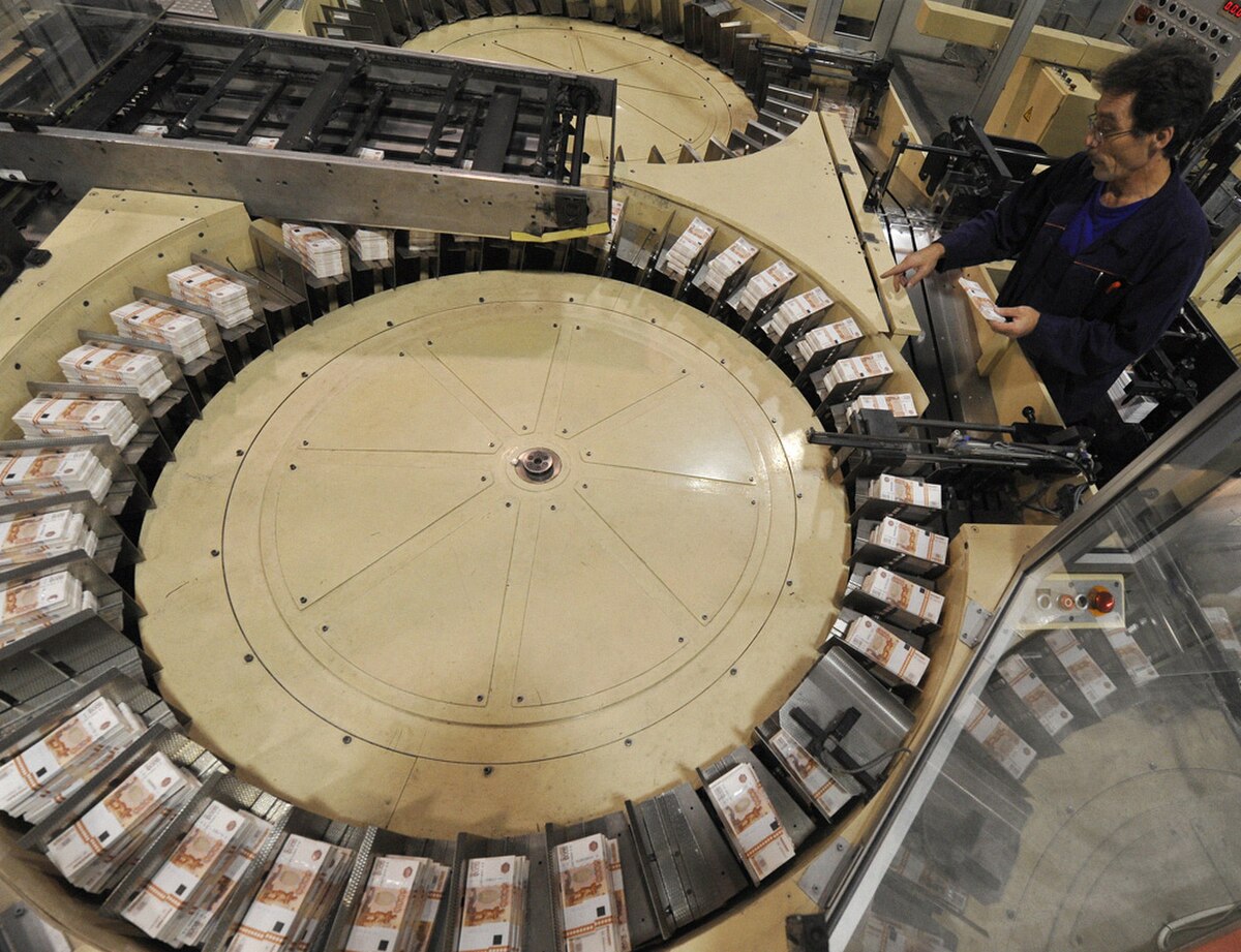 An overhead view of a large industrial machine at the Goznak factory in Perm, where paper money is being printed and stacked in circular arrays. A worker in a blue uniform is visible on the right, overseeing the process.
