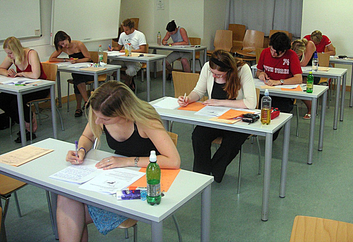 Students are taking a test in a classroom at the University of Vienna, seated individually at desks with papers and writing utensils. The room has a minimalist design with wooden chairs and light-colored walls.