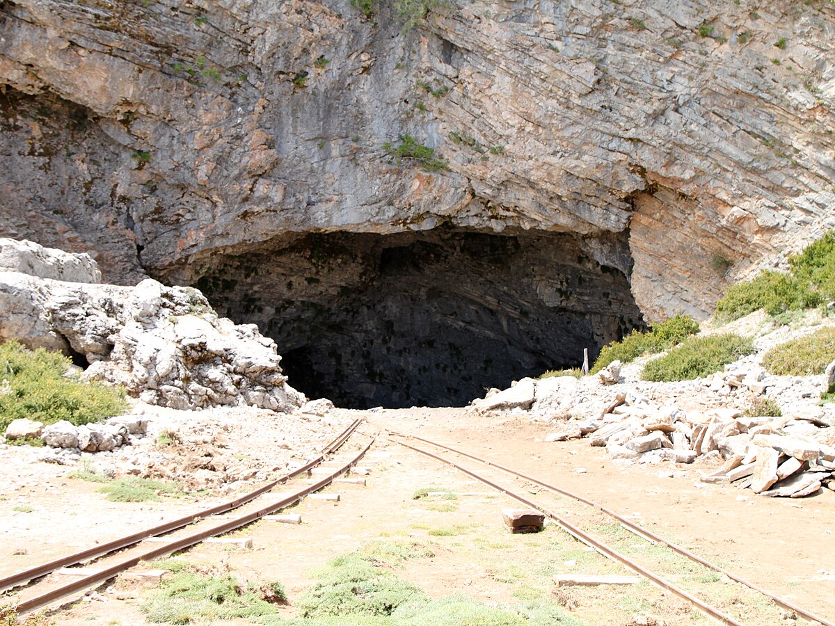 The Cave of Zeus on Mount Ida, Crete, is a large, dark cave entrance at the base of a rocky mountain, with two parallel tracks leading into it. The surrounding area is stony and sparsely vegetated.
