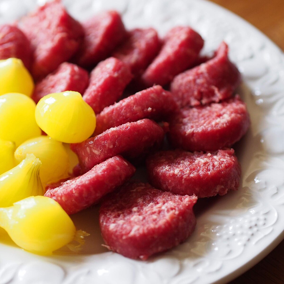 A white plate holds slices of Osseworst, a Dutch beef sausage, alongside small yellow Amsterdamse uitjes (pickled onions).