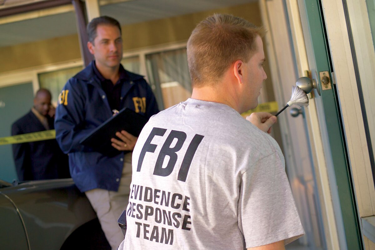 An FBI Evidence Response Team member dusts a doorknob for fingerprints, while another FBI agent in a blue jacket holds a folder and observes. A third person is visible in the background behind yellow crime scene tape.