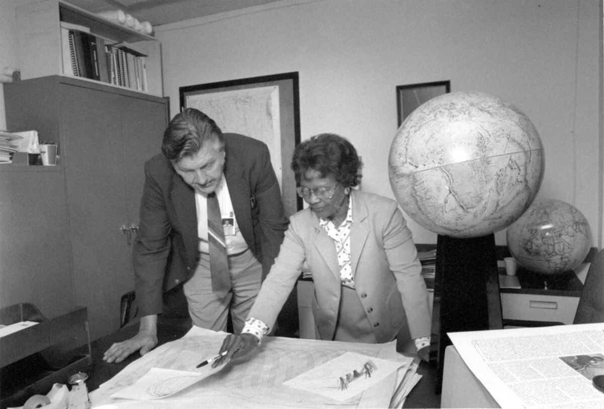 Gladys West and Sam Smith looking over data from the Global Positioning System at Dahlgren in 1985. They are standing over a large map or chart spread across a desk, with two globes visible in the background.