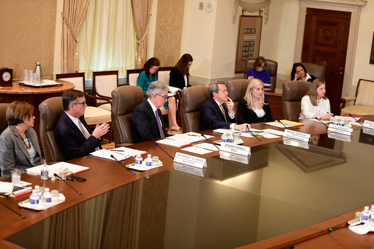 A meeting of the Federal Reserve Board of Governors in April 2019, with several individuals seated around a large wooden conference table. Attendees include men and women in business attire, with documents and water bottles on the table.