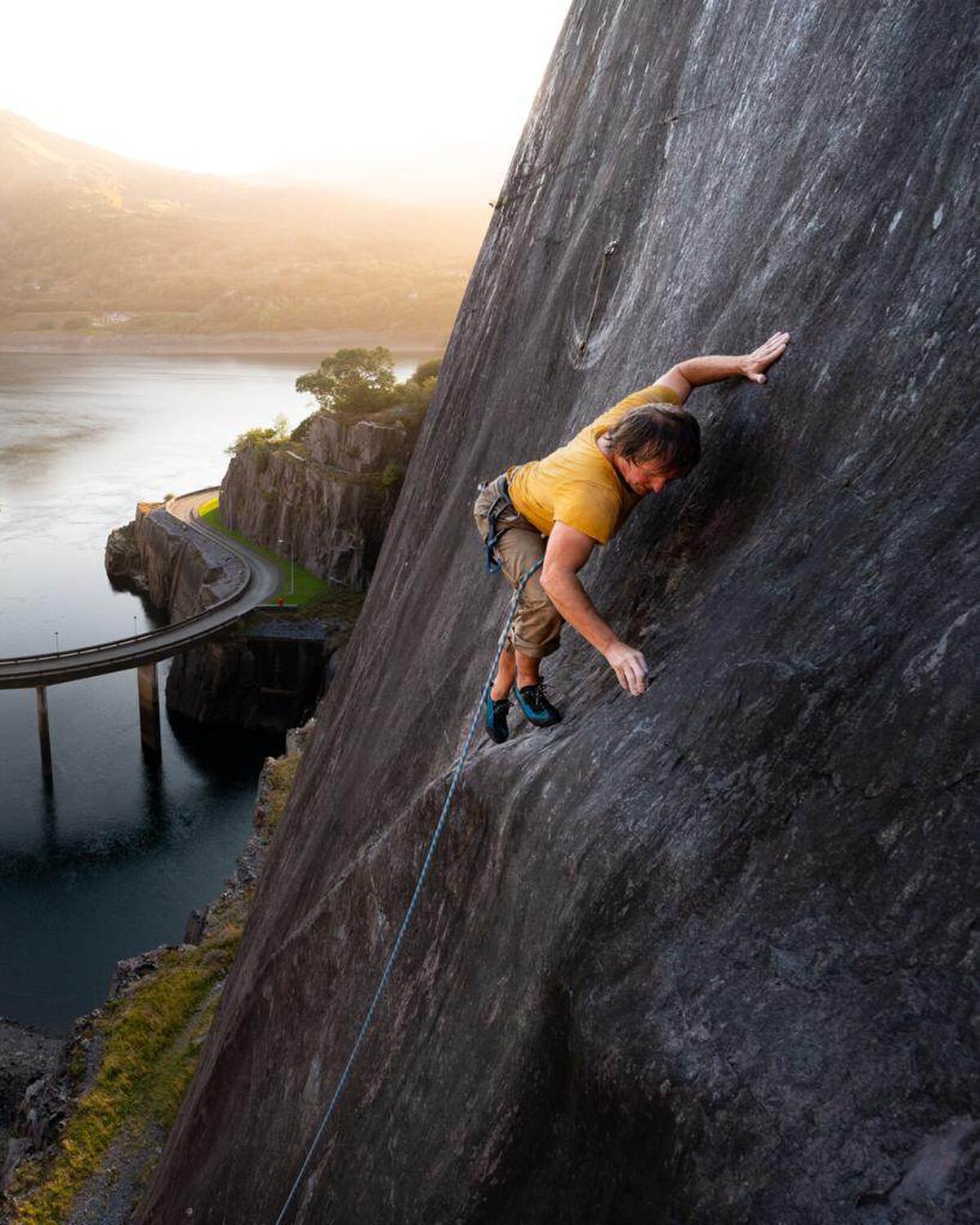Johnny Dawes is shown rock climbing on "Poetry Pink," a route on a large, smooth rock face, with a body of water and a bridge visible in the background. He is wearing a yellow shirt and khaki shorts, with a climbing rope attached to his harness.