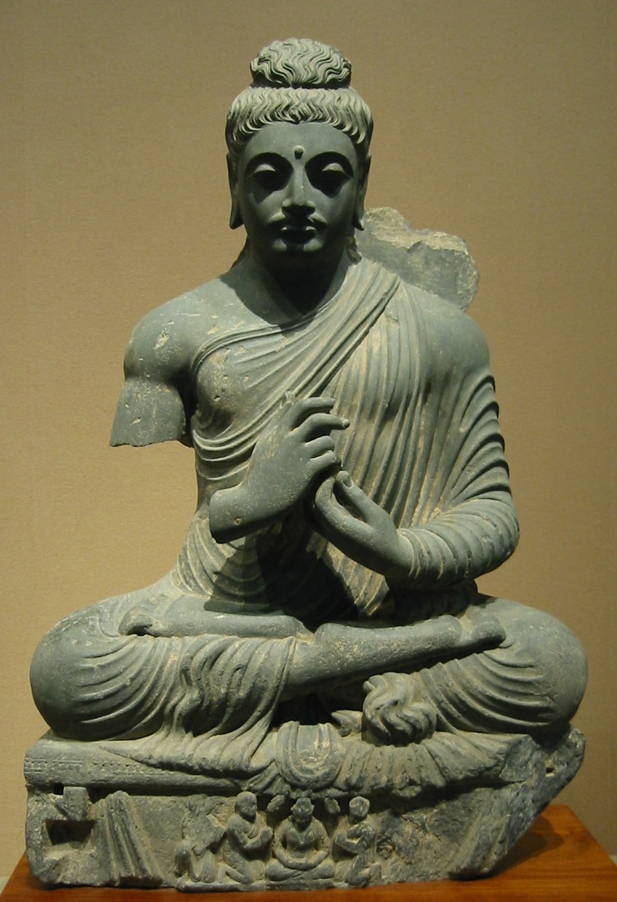 A stone sculpture depicts a seated Buddha with his hands in a teaching mudra. The Buddha is shown with a serene expression, elongated earlobes, and a topknot, with a relief carving of smaller figures at the base.