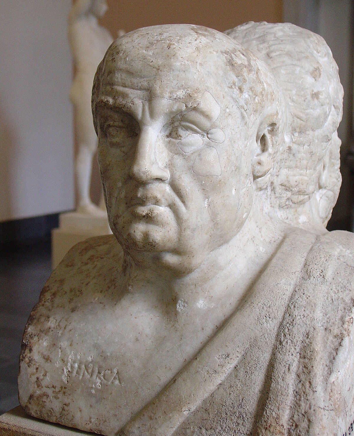 Marble bust of Seneca, with the name "SENECA" carved into the base. The bust shows a man with a receding hairline and a serious expression, with some damage visible on the right side of the face.