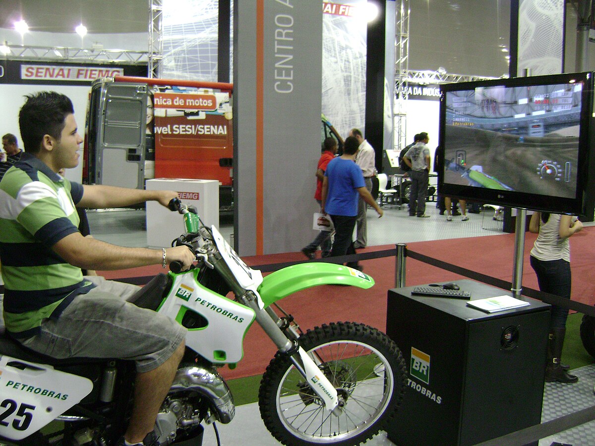 A person sits on a motorcycle simulator, playing a racing video game displayed on a large screen. The motorcycle is branded "PETROBRAS" and has the number "25" on its side, at the Bienal do Automóvel exhibition in Belo Horizonte, Brazil.